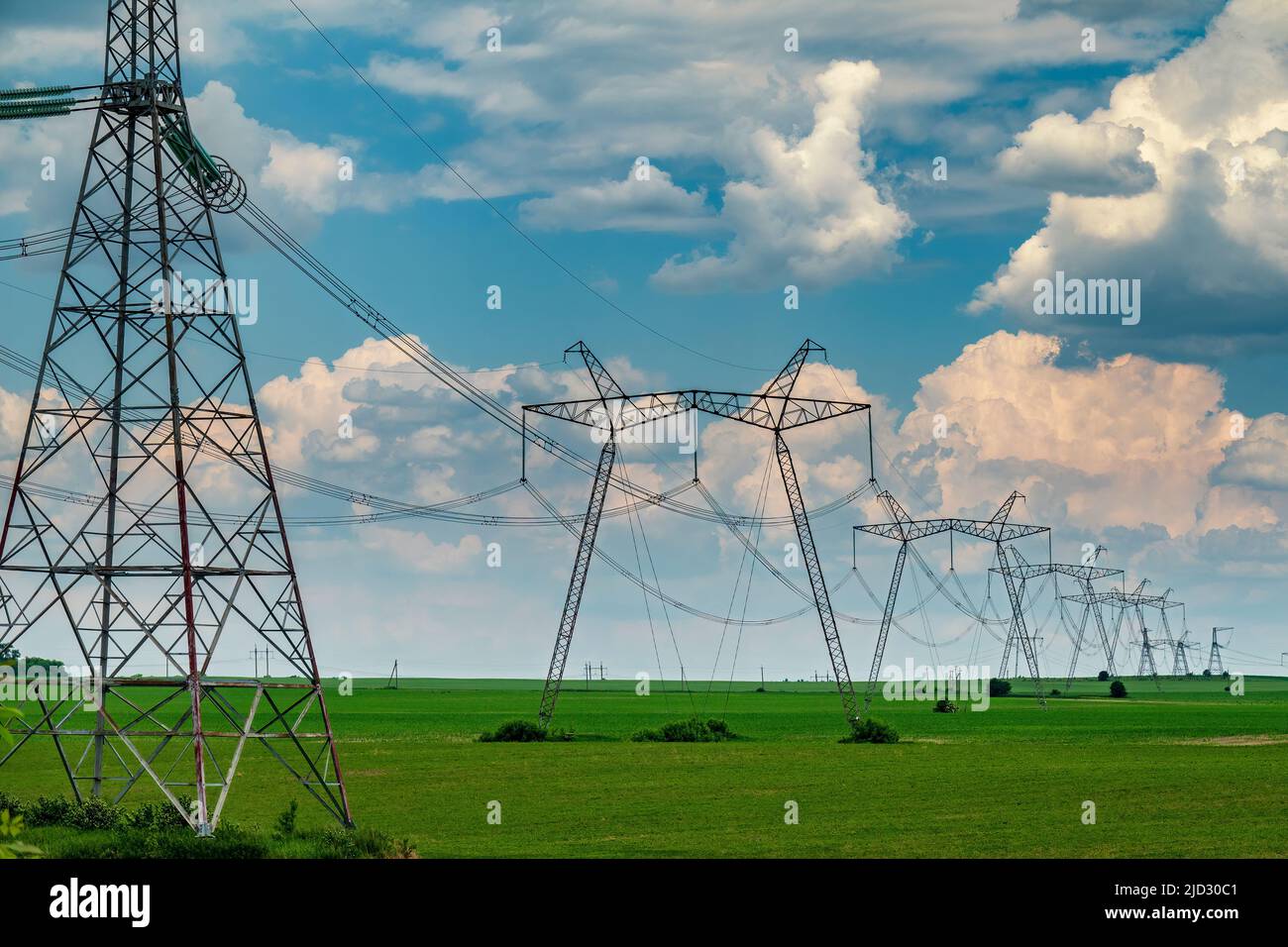 Fila di piloni di supporto della linea di alimentazione sul campo coltivato verde con cielo nuvoloso. Apparecchiature per il passaggio di energia. Foto Stock