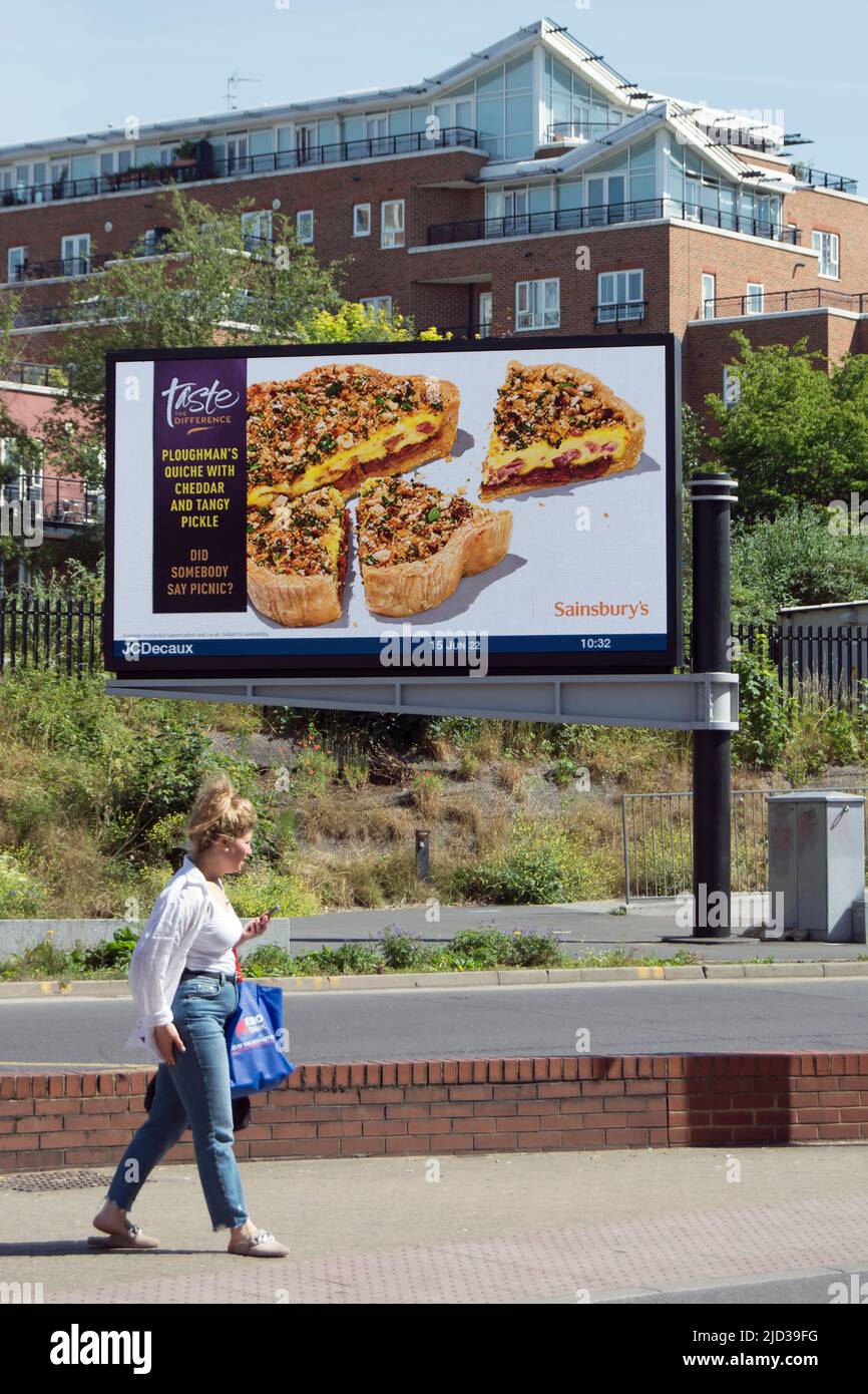 una donna attende di attraversare la strada sotto un cartellone digitale che fa pubblicità alla quiche del plowman di sainsbury, a kingston upon thames, surrey, inghilterra Foto Stock