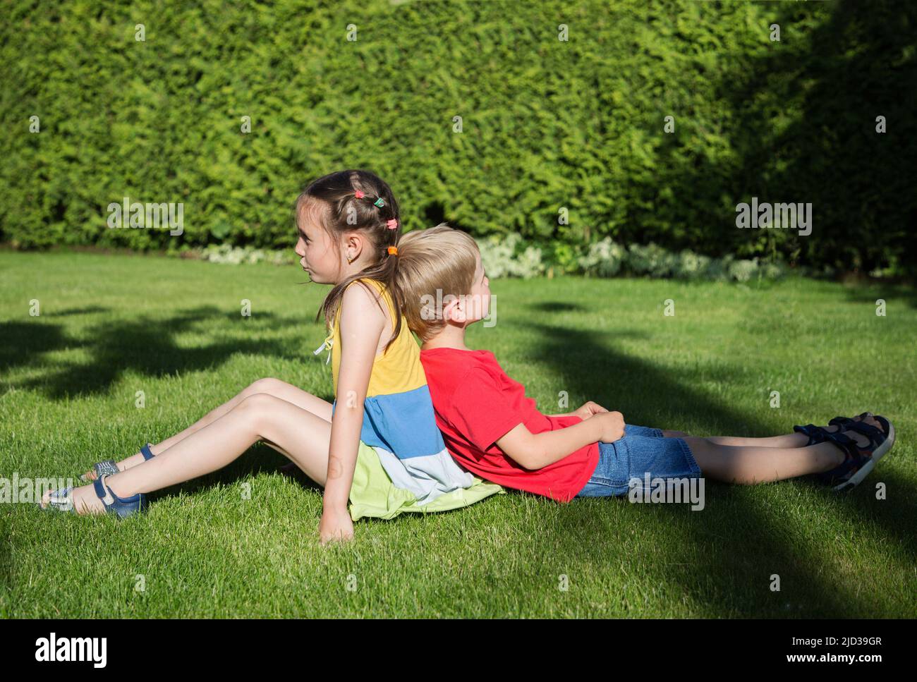 il ragazzo e una ragazza di 6-7 anni sono seduti di nuovo sull'erba in giardino, e parlano e giocano nel verde del parco soleggiato. Amicizia, infanzia, rel Foto Stock