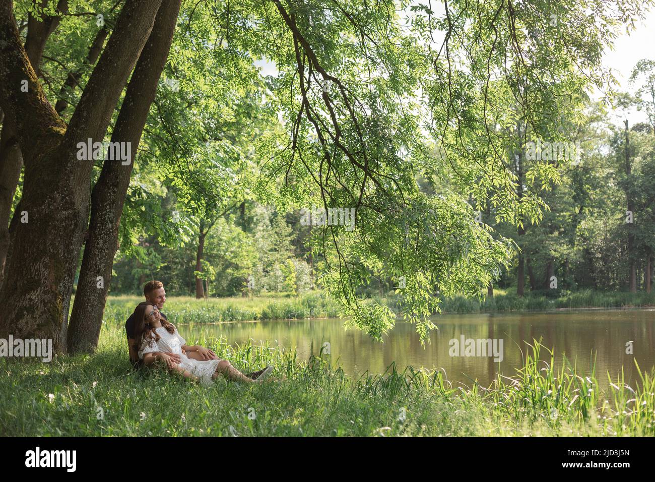 Giovane coppia che si innamorano seduto sull'erba sotto l'albero vicino al lago nel parco, guardando l'un l'altro. Giovane uomo e donna Foto Stock