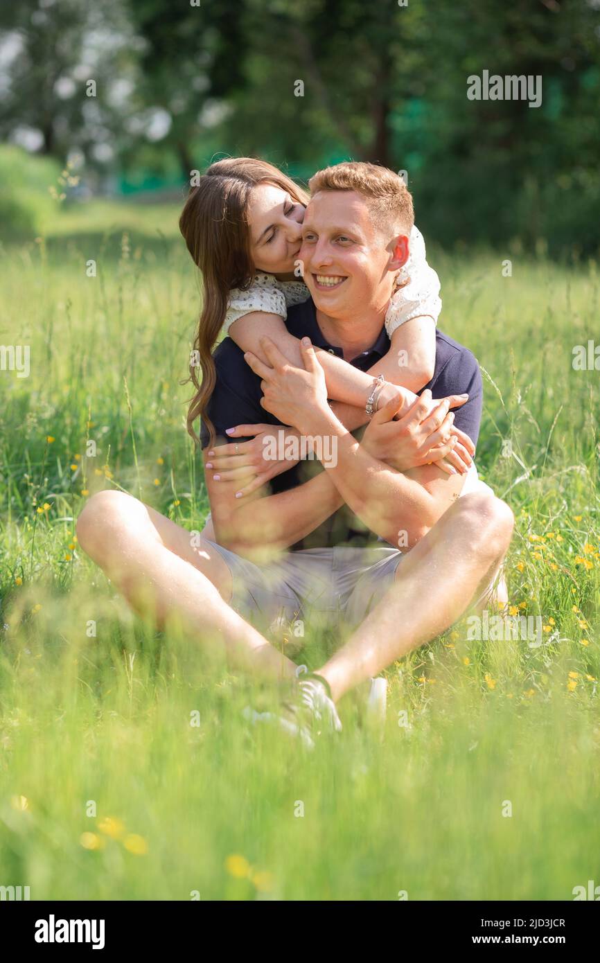 Coppia giovane carina seduta sull'erba, baciare. Giovane uomo e donna che si godono giorno d'estate. Gente felice nell'amore, luna di miele Foto Stock