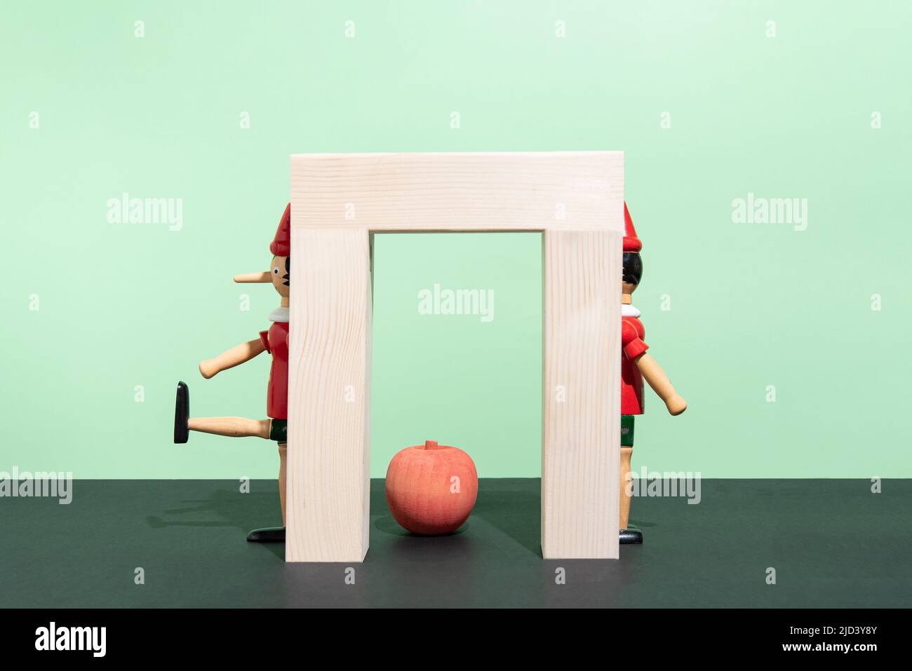 Burattino di legno dietro un cancello di legno e mela su sfondo verde e nero. Concetto minimo creativo. Layout rettangolo con spazio di copia Foto Stock