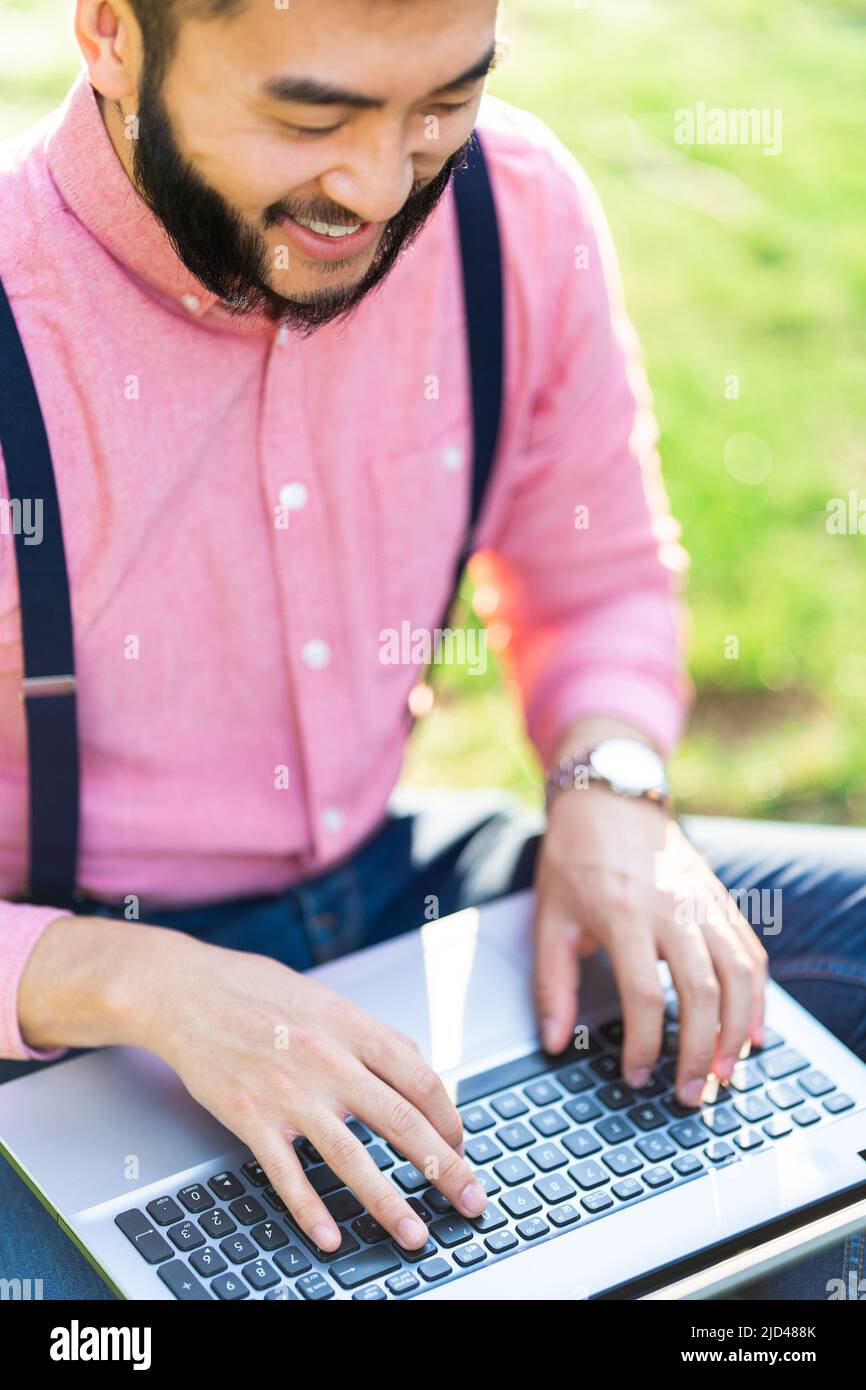 Uomo asiatico moderno che lavora con un computer portatile in un parco Foto Stock