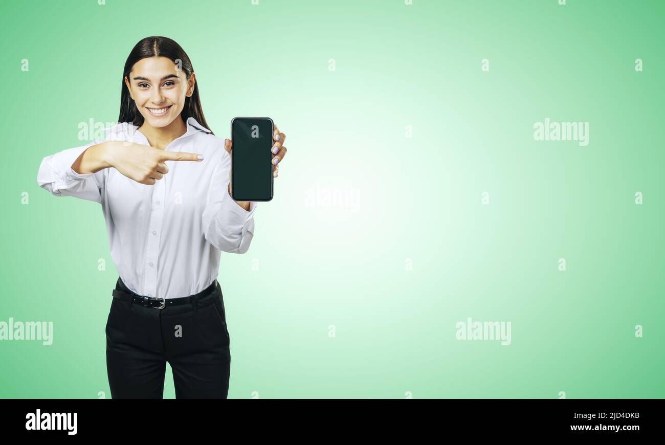 Concetto di applicazione mobile con ragazza felice in camicia bianca mostrando moderno cellulare con schermo vuoto su sfondo astratto verde chiaro con posto per Foto Stock