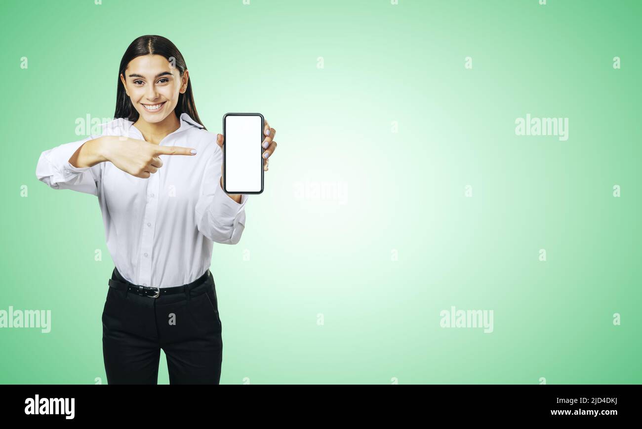 Concetto di applicazione mobile con donna d'affari sorridente in camicia bianca che mostra il cellulare moderno con schermo bianco vuoto su sfondo verde astratto wit Foto Stock