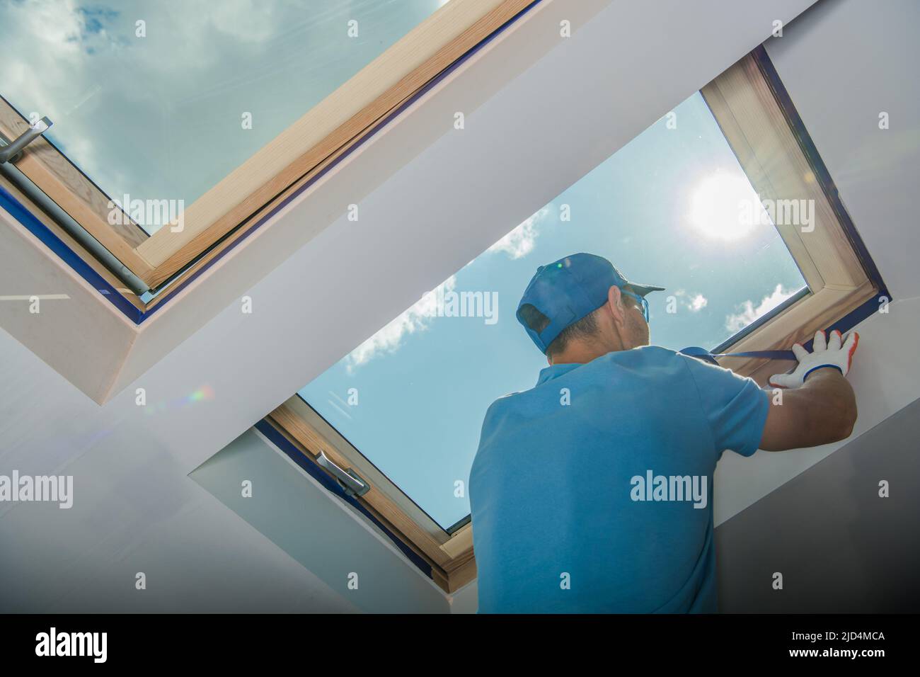 Lavoratore caucasico maschio in Blue Outfit preparazione finestra lucernario prima di dipingere le pareti bianche nella stanza. Tema lavori di ristrutturazione. Foto Stock