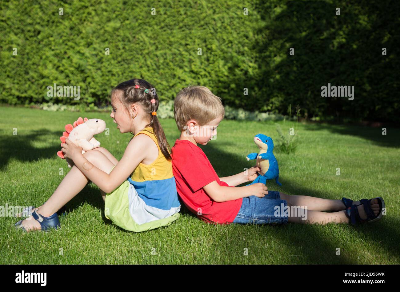 ragazzo e una ragazza di 6-7 anni sono seduti di nuovo sull'erba in giardino. giocare a giochi di fantasia con lussuosi dinosauri. Amicizia, infanzia, s Foto Stock