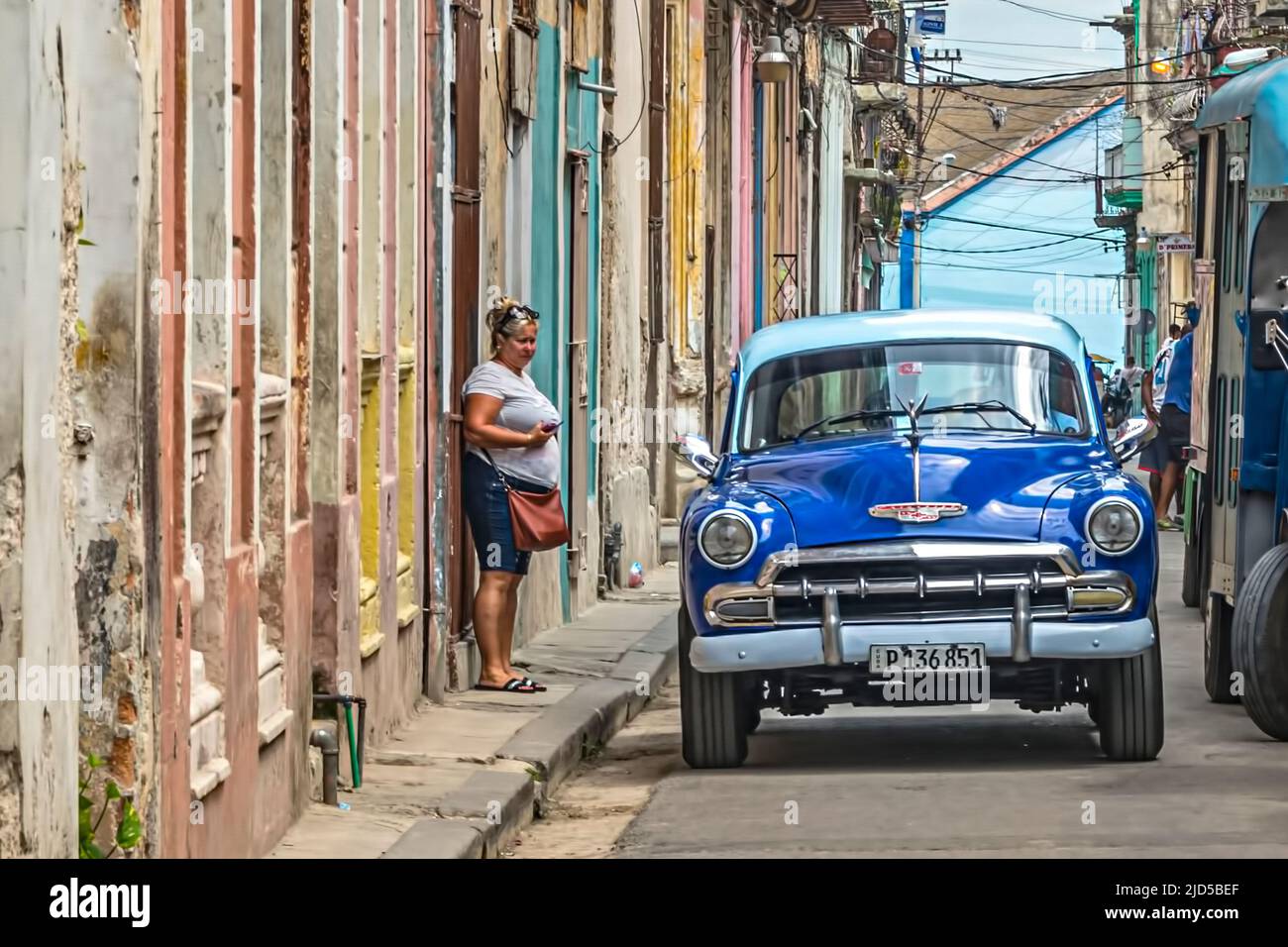 Signora cubana in una strada con un'auto d'epoca blu che passa a Old Havana, Cuba Foto Stock
