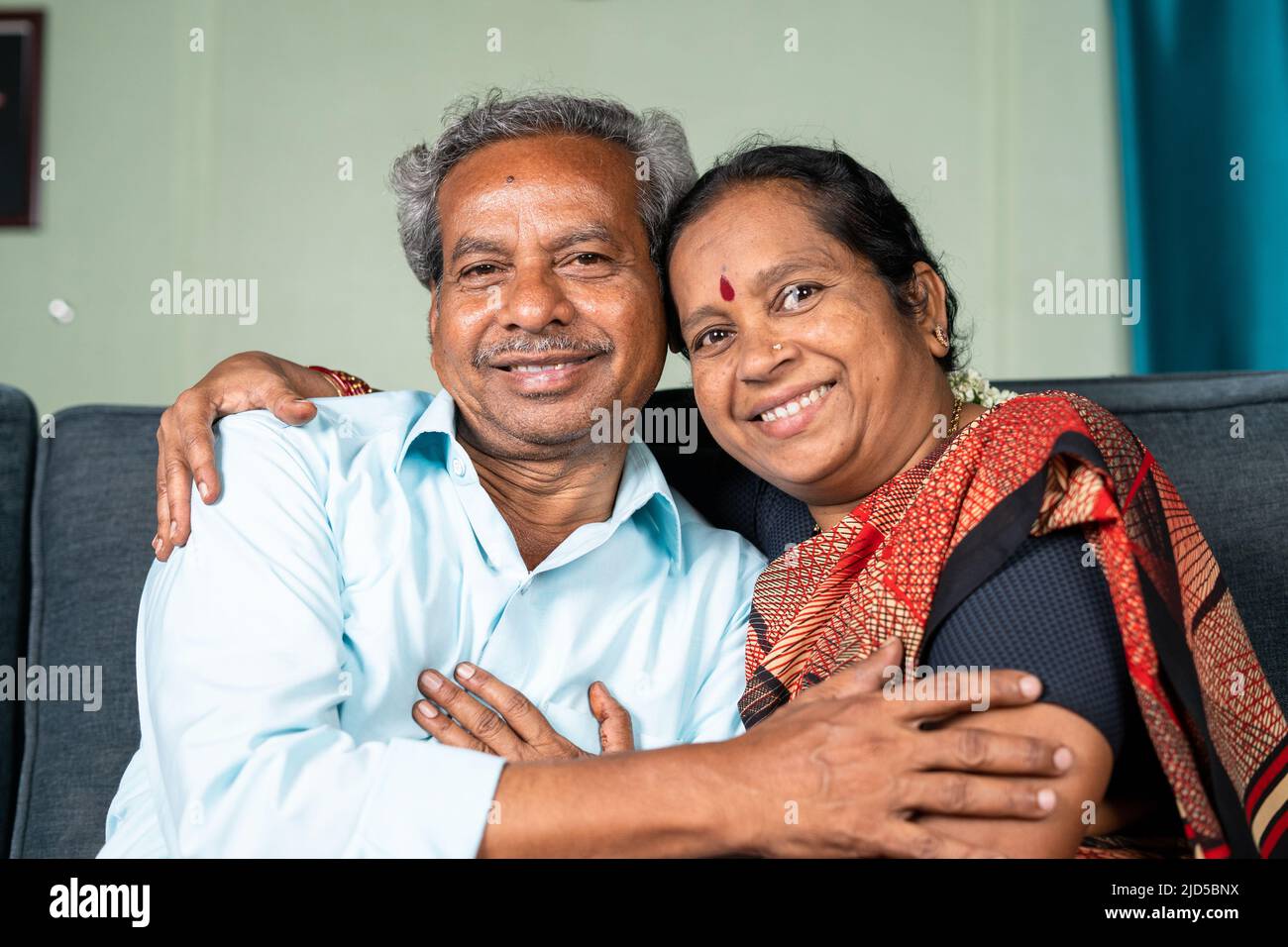 Felice sorridente coppia indiana senior abbracciandosi a vicenda guardando la macchina fotografica mentre sul divano - concetto di legame familiare, relax e felicità Foto Stock