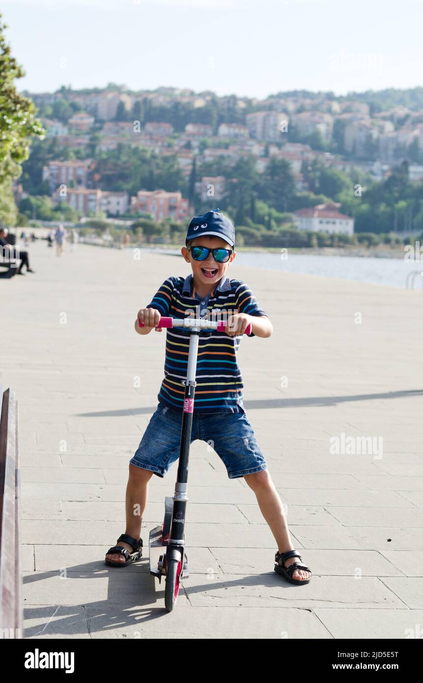 concetto di vacanza. bambino che guida scooter, a piedi nella città vecchia, strada. Risate in una giornata estiva di sole. Divertirsi Foto Stock