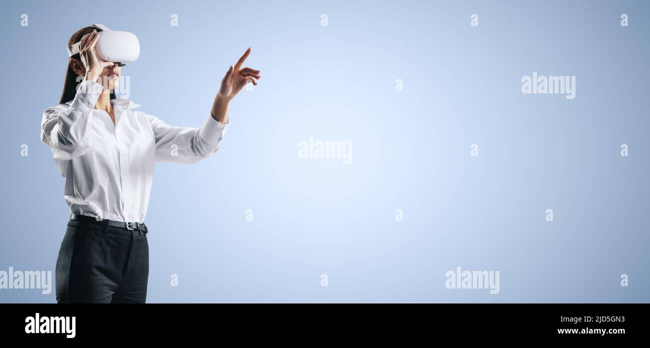 La giovane donna che lavora in un ambiente di realtà virtuale con occhiali VR, preme con il dito sull'interfaccia virtuale sullo sfondo blu astratto. Innovazione e f Foto Stock