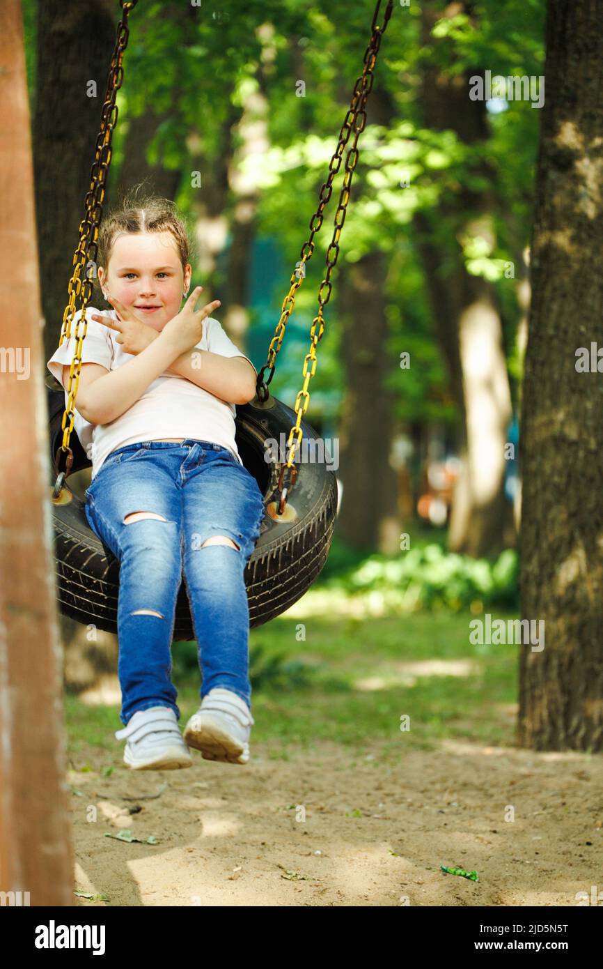 Piccola ragazza fredda che oscilla su ruota di gomma dell'automobile con le braccia incrociate nel parco giochi nel parco verde. Vacanze estive in campeggio, centro turistico. Giocare all'aperto Foto Stock