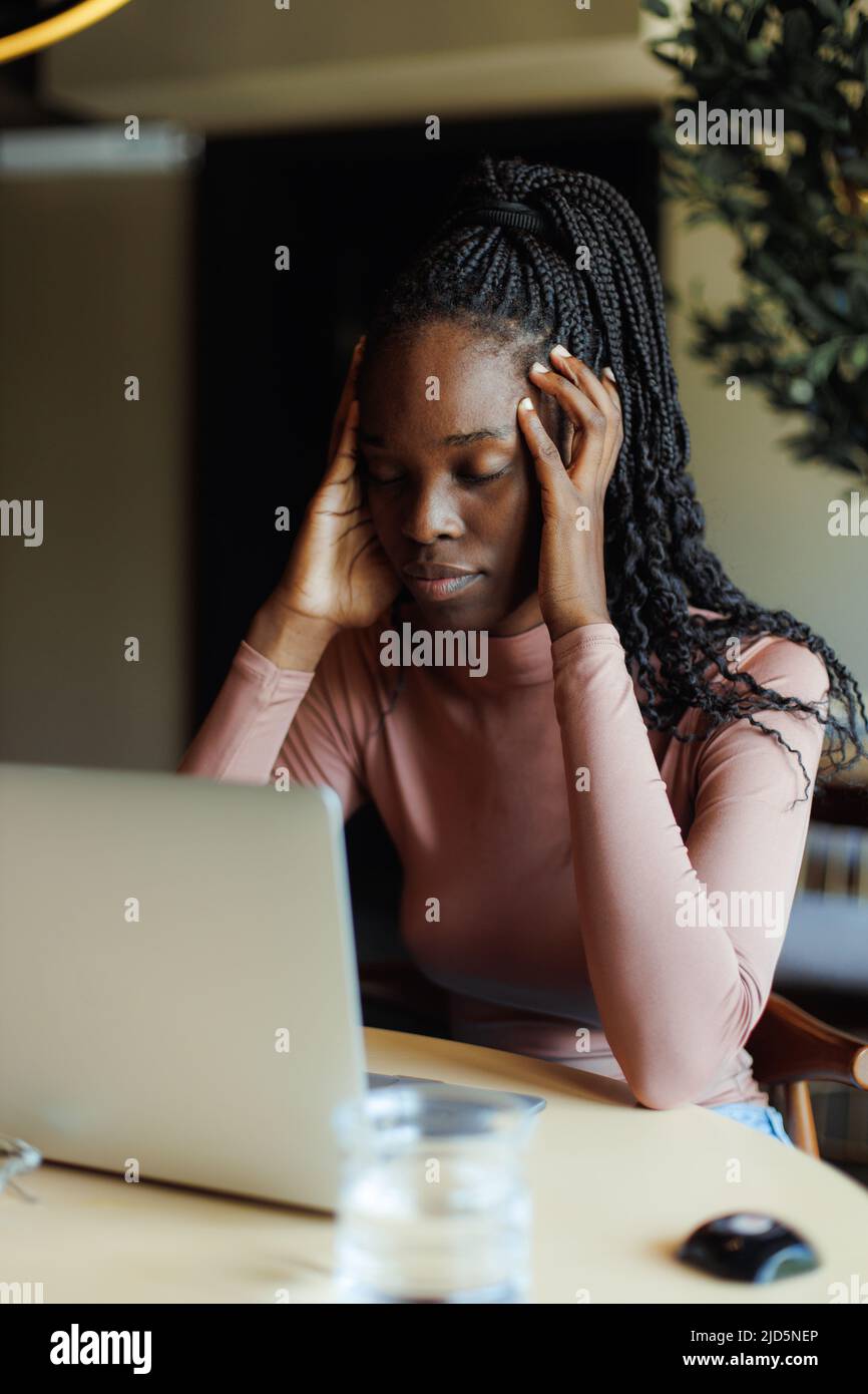 Giovane afroamericana che pensa con occhi chiusi e lavora online su un portatile in una caffetteria. Ragazza nera che lavora a distanza, freelance, confortevole Foto Stock