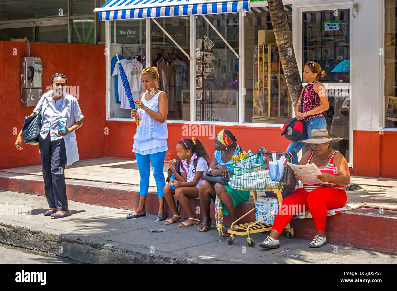 Donne colorate vestite in attesa alla fermata dell'autobus con carrello per lo shopping a Santiago de Cuba, Cuba Foto Stock