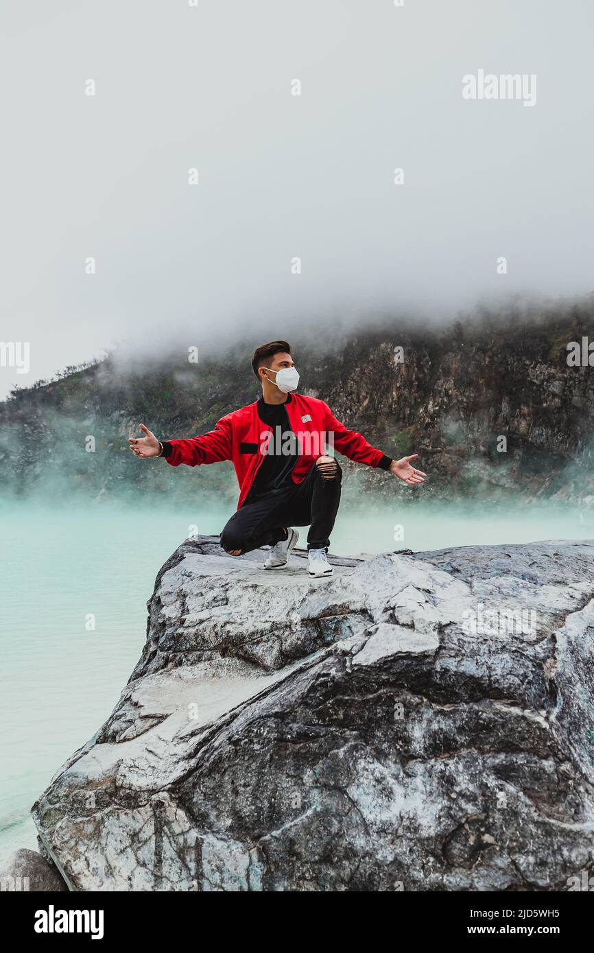 giovane uomo in bomber rosso inginocchiato sulla roccia al lago di kawah putih sulfer Foto Stock