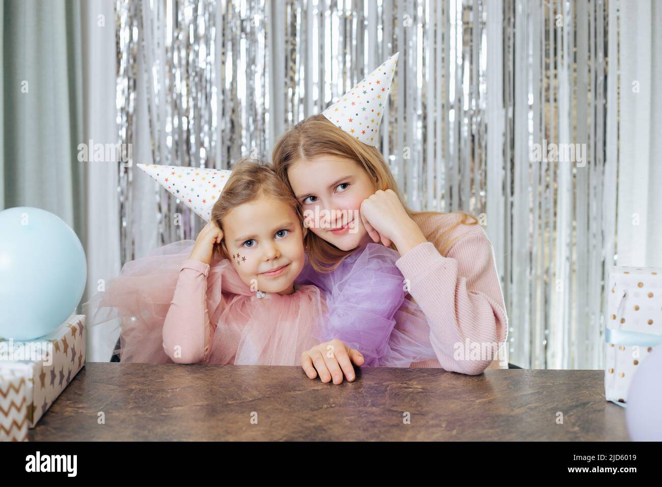Ritratto di due belle sorelle che celebrano il loro compleanno. Le ragazze in cappelli da festa e abiti da festa si siedono allo stesso modo, con le loro guance propped in su con Foto Stock