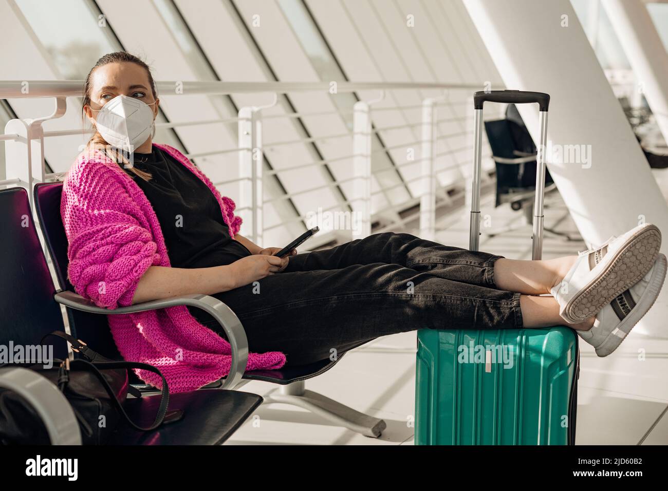 Ritratto di donna pantaloni neri, cardigan rosa, bianco maschera usa e getta, seduta, gambe stretching, tenere in mano lo smartphone. Foto Stock