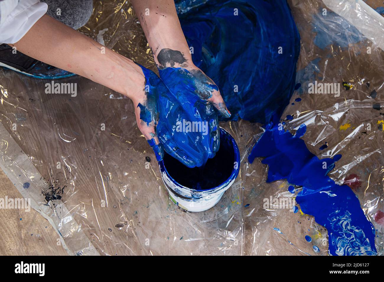 Foto ritagliata di mani piegate di donna macchiate con vernice blu sopra secchio su pellicola trasparente ricoperta da spruzzi. Foto Stock