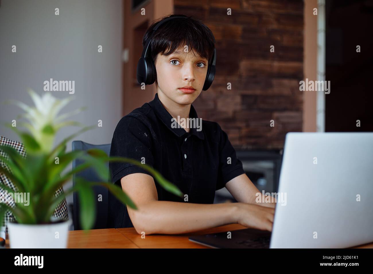 Ritratto di ragazzo adolescente con capelli corti e scuri indossando le cuffie, seduto alla scrivania a casa, digitando sul portatile, posando. Foto Stock