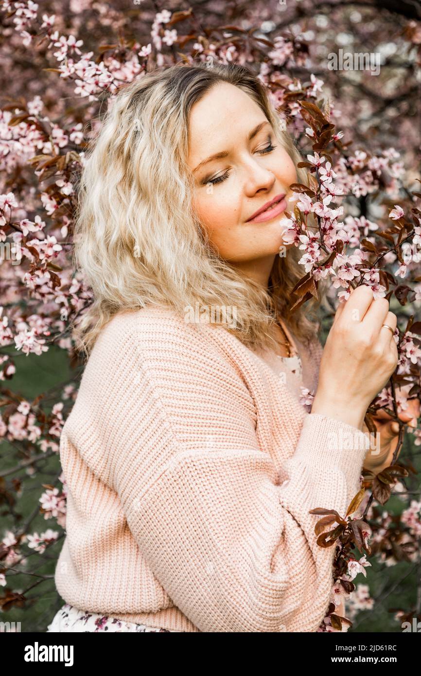 Giovane donna con occhi chiusi in piedi vicino a un albero di ciliegio in fiore, che tiene ramo nel parco, fioriture di fiore bianco. Foto Stock