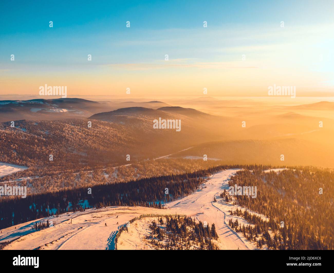 Pista da sci in una zona montagnosa nella foresta al tramonto, vista aerea. Sciatori e snowboarder si snollano lungo la montagna con viste mozzafiato della natura. Foto Stock