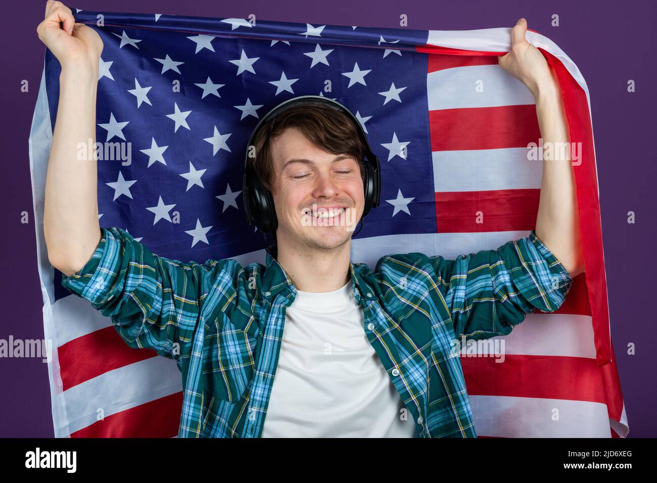 L'uomo felice in una camicia tiene una bandiera americana e ascolta musica con le cuffie su sfondo viola. La celebrazione della Giornata dell'Indipendenza il 4 luglio. Foto Stock