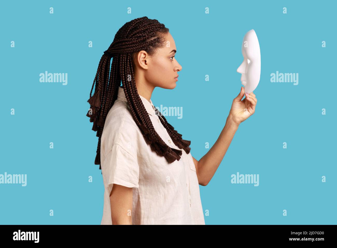 Vista laterale della donna seria sicura con i dreadlocks che tengono e guardano la maschera bianca con lo sguardo attento, cercando di capire la personalità nascondente. Studio interno girato isolato su sfondo blu. Foto Stock