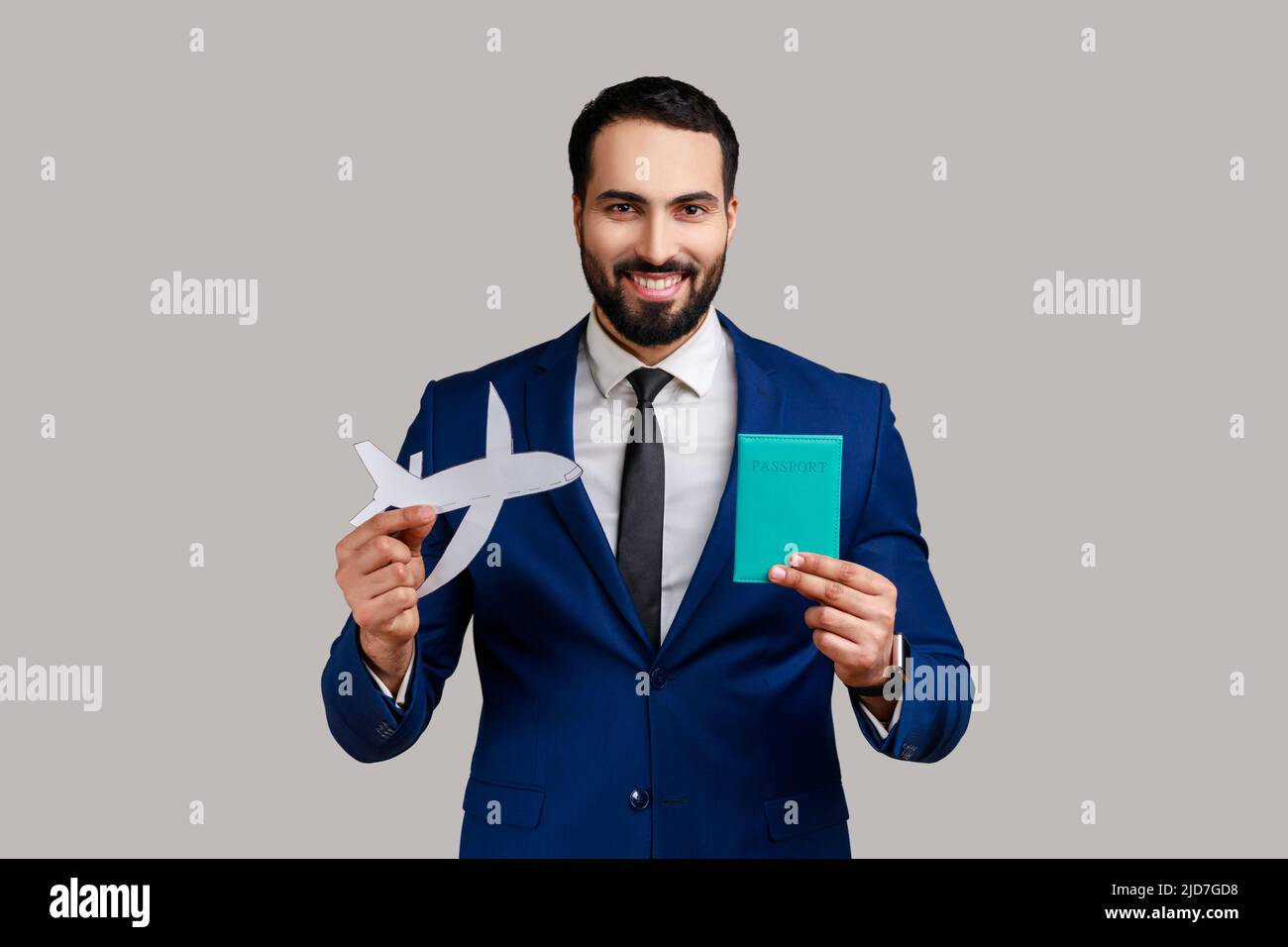 Ritratto di positivo felice uomo barbuto sorridendo e mostrando passaporto e aereo di carta a macchina fotografica, turismo, indossando tuta di stile ufficiale. Studio interno girato isolato su sfondo grigio. Foto Stock