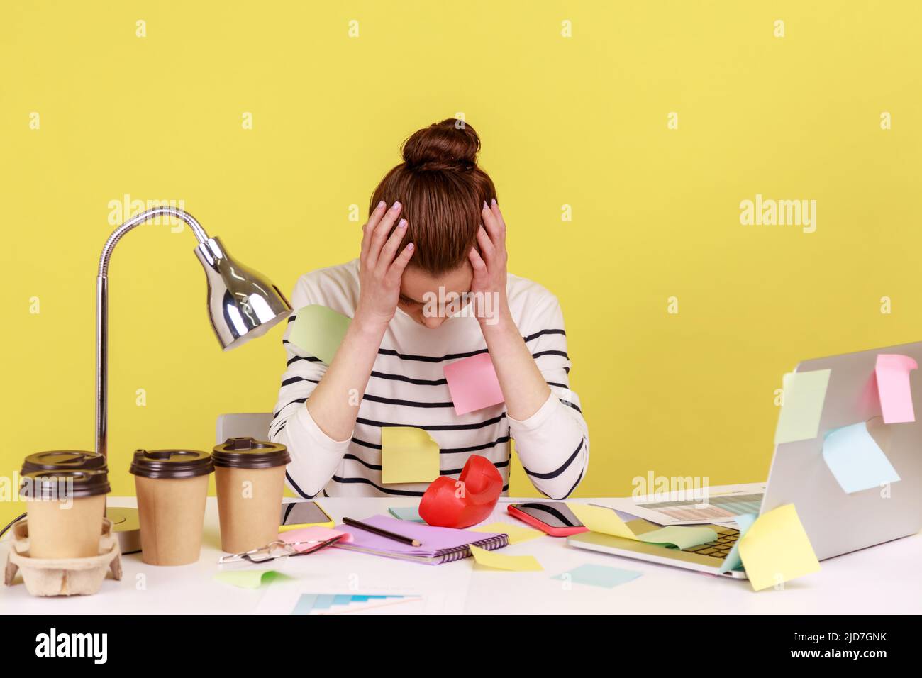 Stanco esausto lavoratore di ufficio donna sovraccaricato coperto di note appiccicose, testa stringente, pensiero disperato, mal di testa soffrendo. Studio interno girato isolato su sfondo giallo. Foto Stock