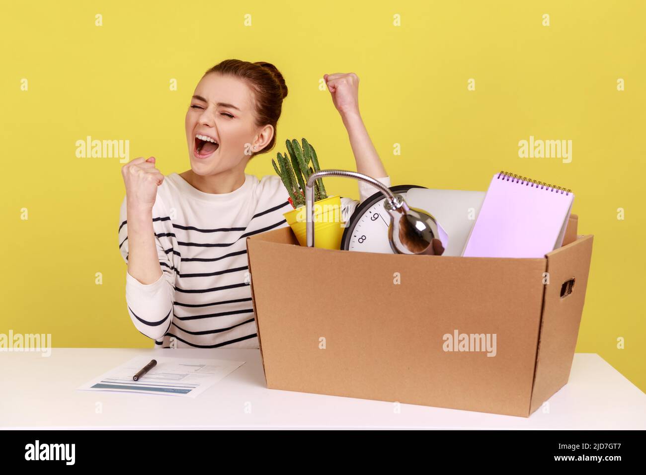 Ritratto di felice donna eccitata seduta sul tavolo con la scatola con il suo personale roba, gioendo per ottenere nuovo lavoro, esprimendo le emozioni positive. Studio interno girato isolato su sfondo giallo. Foto Stock