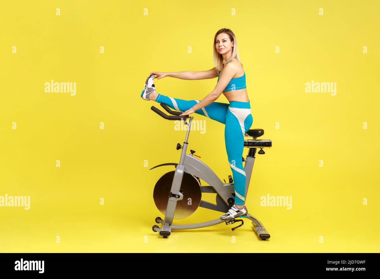 Ritratto a tutta lunghezza di bella atletica donna che stretching gamba prima di allenarsi in bici esercizio, guardare via, indossando abbigliamento sportivo blu. Studio interno girato isolato su sfondo giallo. Foto Stock