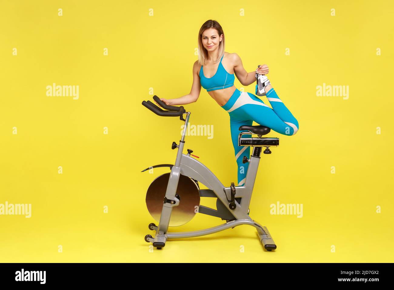 Ritratto completo di donna sottile con corpo perfetto in piedi su una gamba della cyclette, guardando la fotocamera, indossando abbigliamento sportivo blu. Studio interno girato isolato su sfondo giallo. Foto Stock