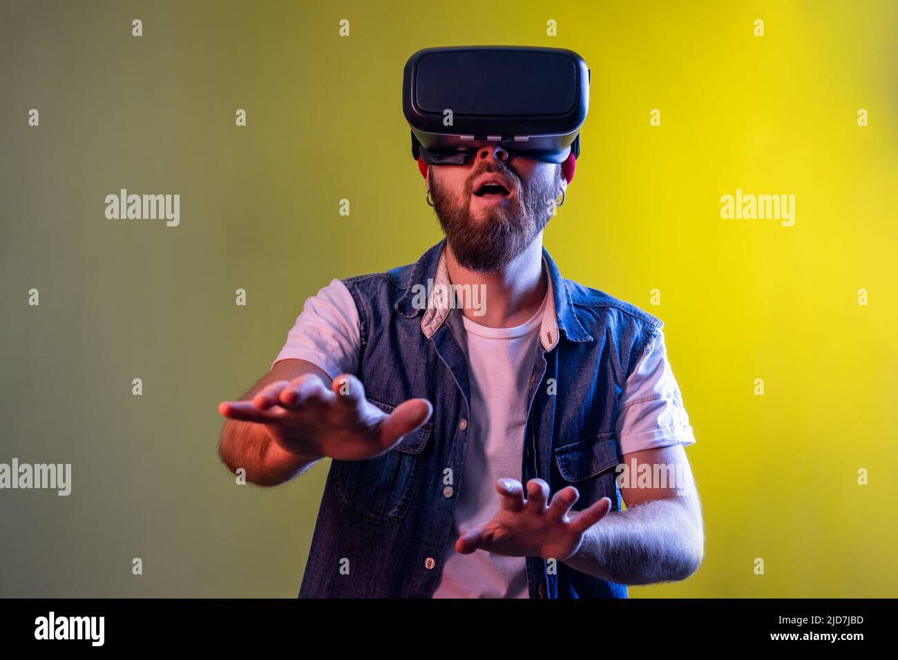 Stupito uomo hipster in piedi in realtà virtuale cuffie, giocare video gioco e cercare di toccare qualcosa, indossare cappello e denim gilet. Studio interno girato isolato su colorato sfondo di luce al neon. Foto Stock