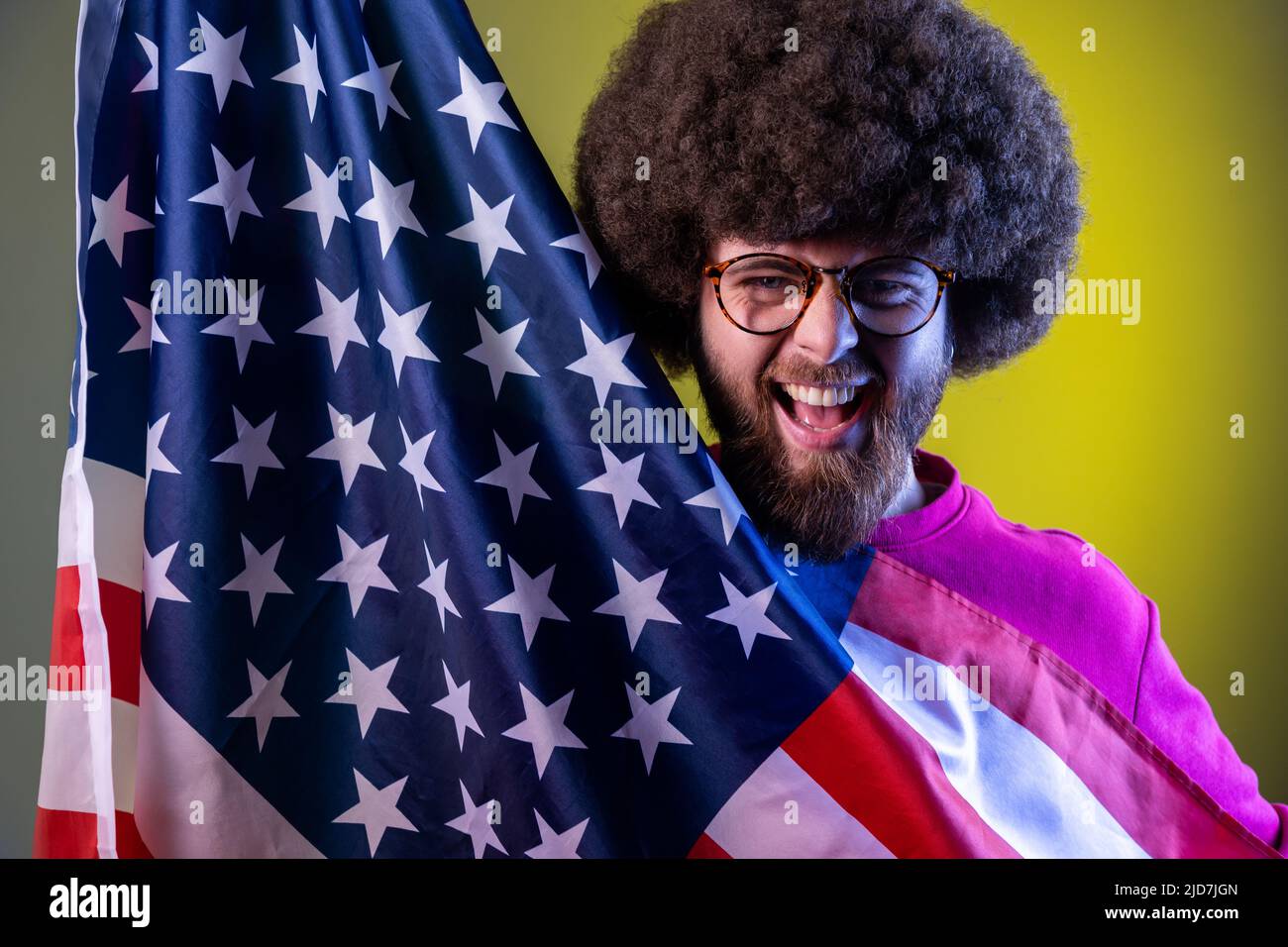 Ritratto di hipster uomo con acconciatura Afro divertirsi, tenere bandiera americana, gioire, celebrare le festività nazionali. Studio interno girato isolato su colorato sfondo di luce al neon. Foto Stock