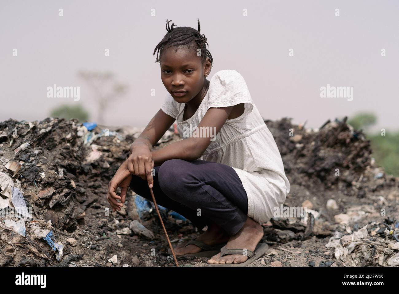 Triste ragazza africana con un aspetto accusatorio inginocchiarsi in mezzo ai rifiuti, contemplando il disastro ambientale del suo paese Foto Stock