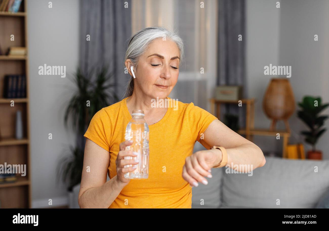 Donna sportiva senior con bottiglia d'acqua che controlla il suo orologio o fitness tracker, che riposa sul divano dopo l'allenamento domestico Foto Stock