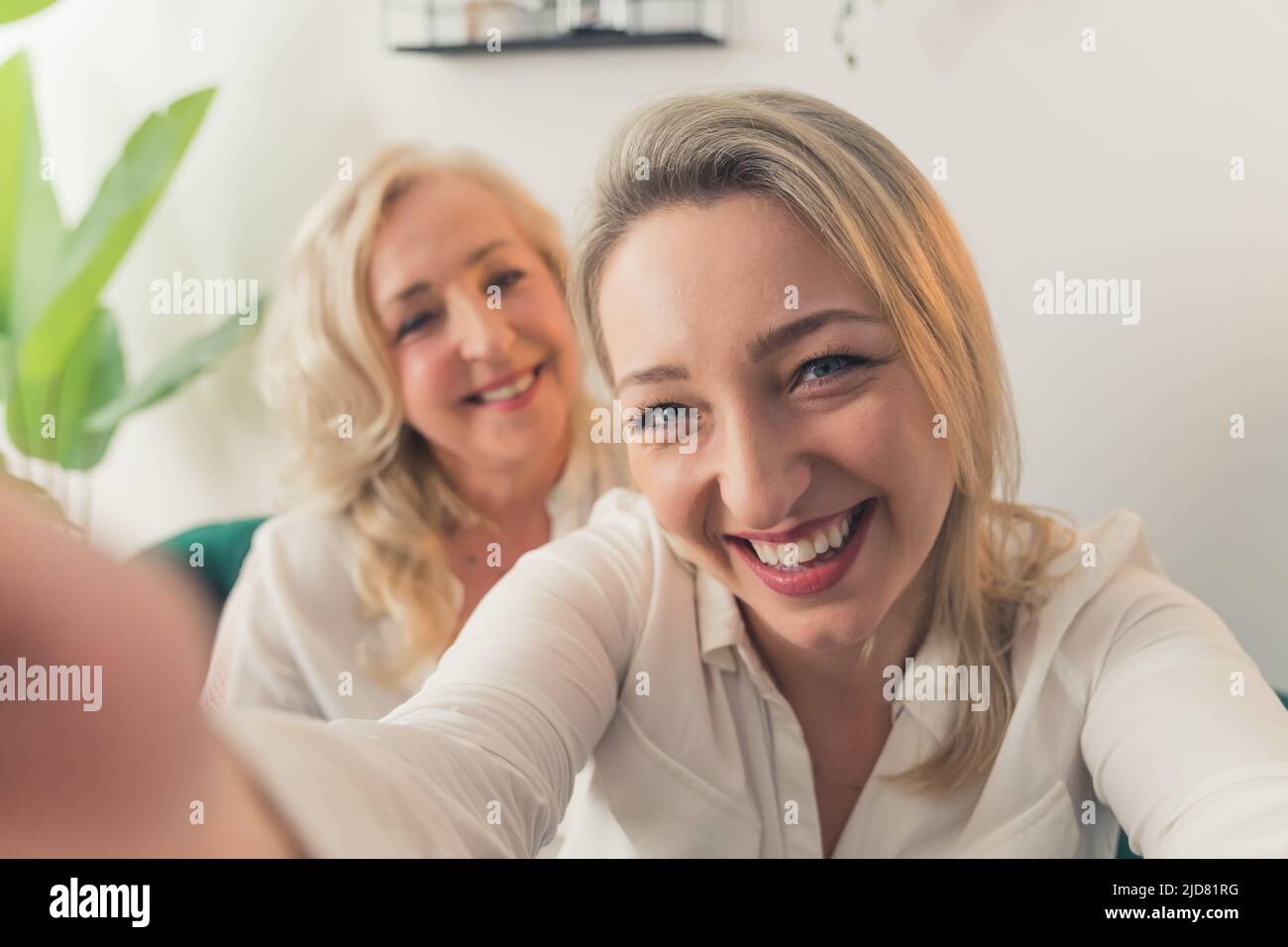 Donna europea di mezza età dai capelli biondi in una camicia bianca che prende un selfie con la sua madre simile in piedi nel backgruond. Scatto al coperto. Foto di alta qualità Foto Stock