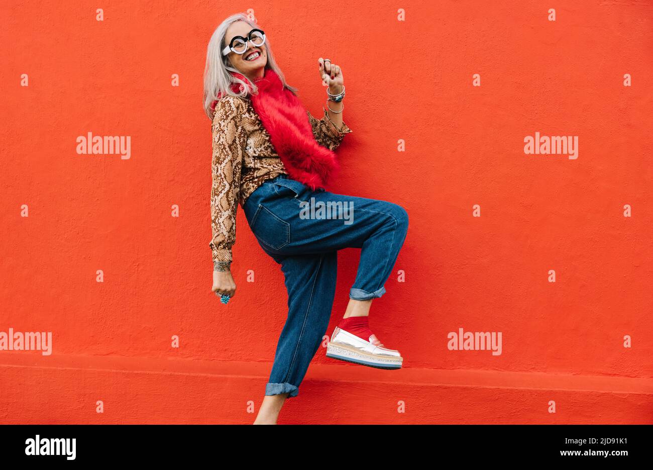 Donna anziana attiva che balla allegro su sfondo rosso. Donna anziana felice sentendosi giovane e vibrante, indossando abiti colorati. Es Foto Stock