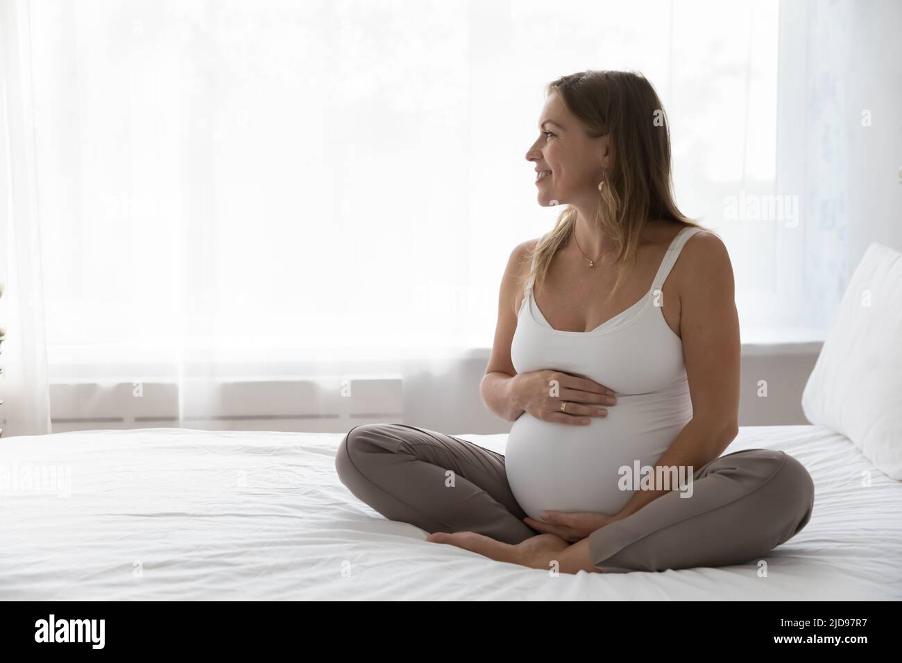 Donna incinta felice seduta sul letto guarda da parte Foto Stock