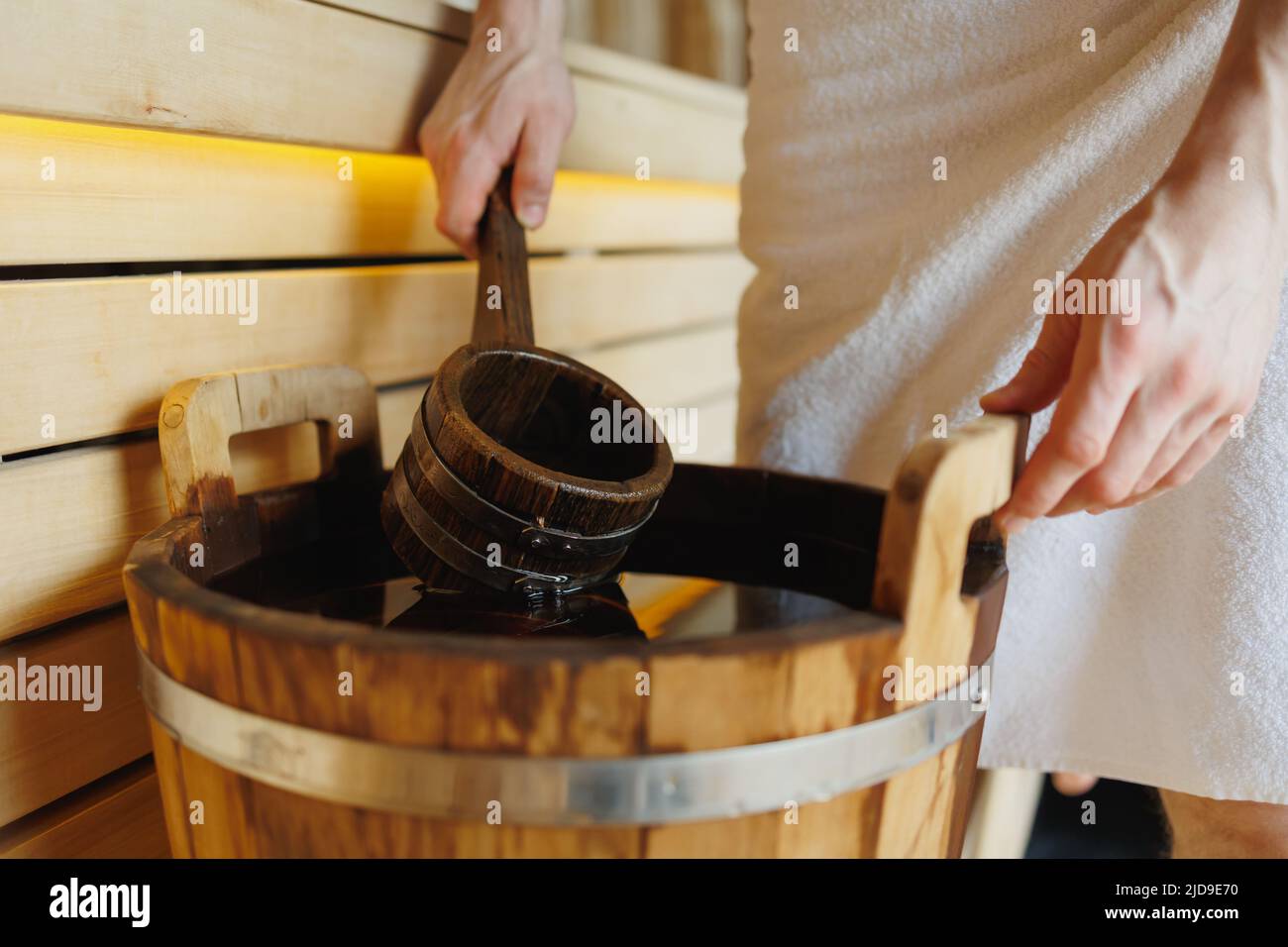Primo piano di un uomo in un asciugamano che raccoglie acqua con un mestolo da un secchio di legno in una sauna Foto Stock