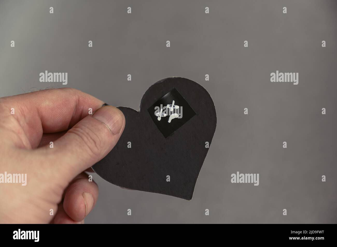 Un uomo che tiene in mano un cuore nero. Un volto triste con occhi incrociati è disegnato in bianco su un cuore di carta. La mano di un uomo di mezza età. Non richiesta Foto Stock