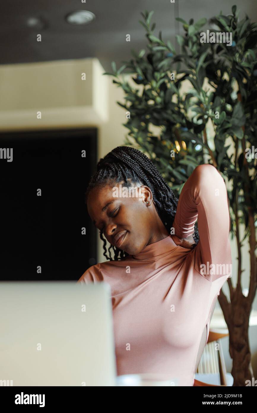 Giovane afroamericana donna stretching collo con occhi chiusi inclinando testa a lato, seduta e lavoro in caffetteria. Ragazza nera che lavora da remoto Foto Stock