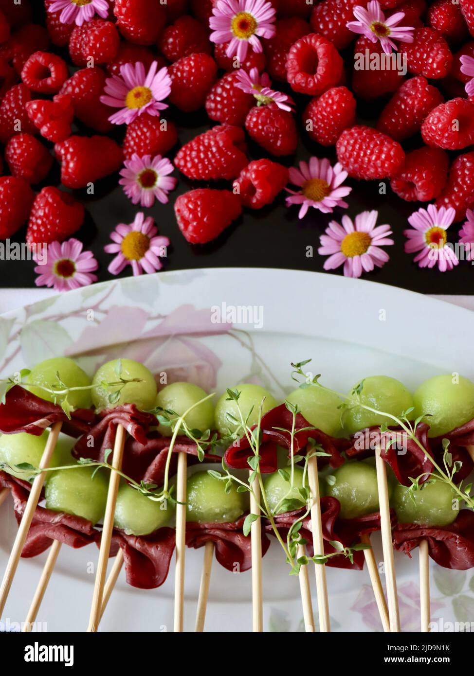 Spiedini di bambù con renna affumicata e melone su vassoio decorato con lamponi e fiori viola. Foto Stock