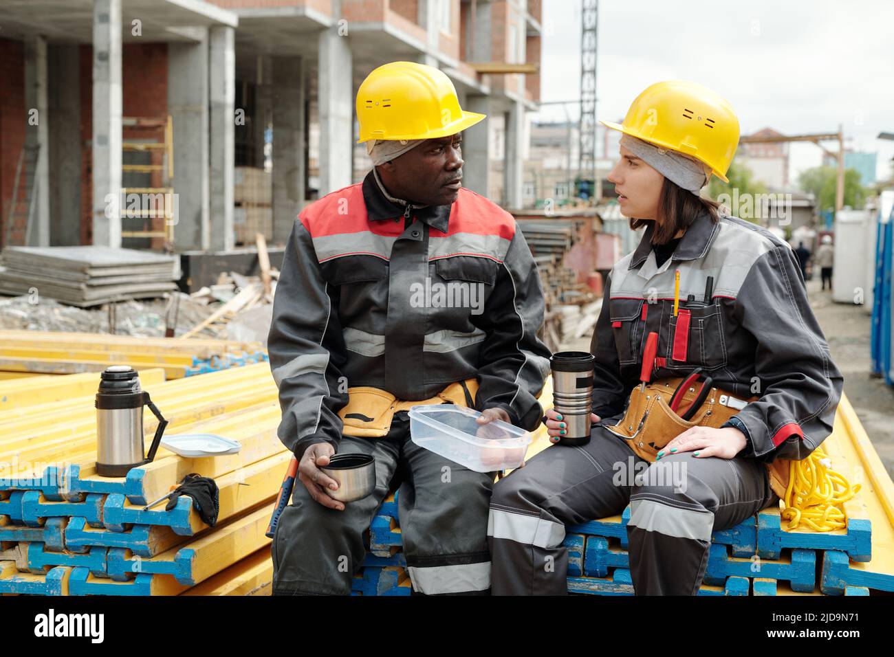 Due ingegneri o costruttori moderni con tazze di tè caldo che hanno parlato in cantiere mentre si siede su una pila di materiali da costruzione gialli Foto Stock