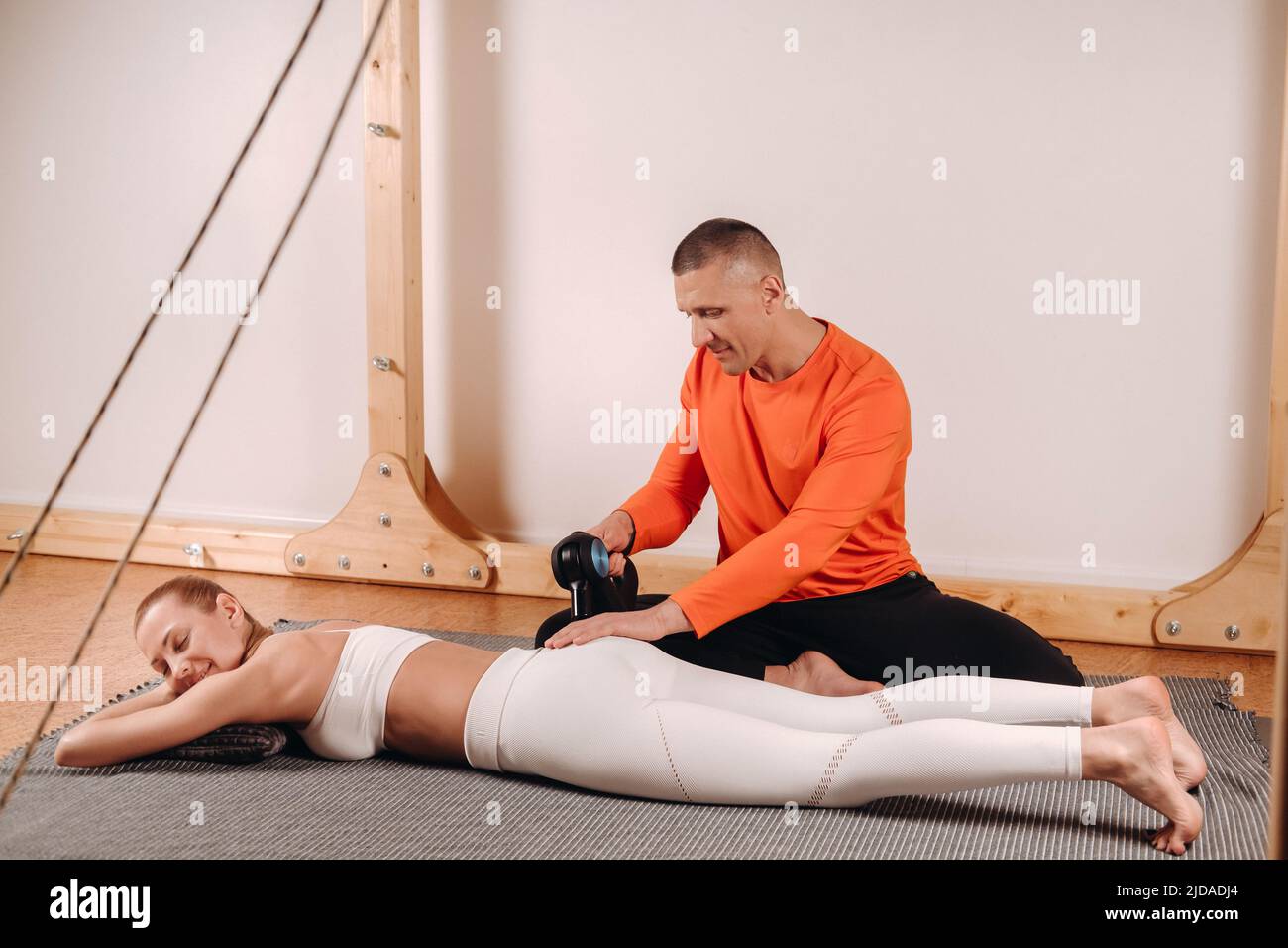 Un massaggiatore maschile massaggia il corpo di una ragazza con un massaggiatore a percussione per preparare un simulatore di stretching. Foto Stock