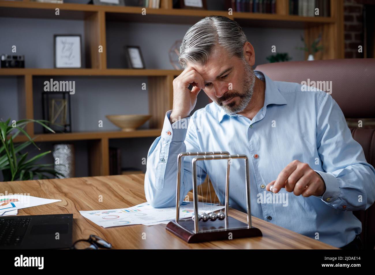 Uomo d'affari stanco seduto alla scrivania e giocando con le palle di bilanciamento della culla di Newton. Annoiato manager rilassarsi dopo il lavoro. Burnout e lavoro in eccesso Foto Stock