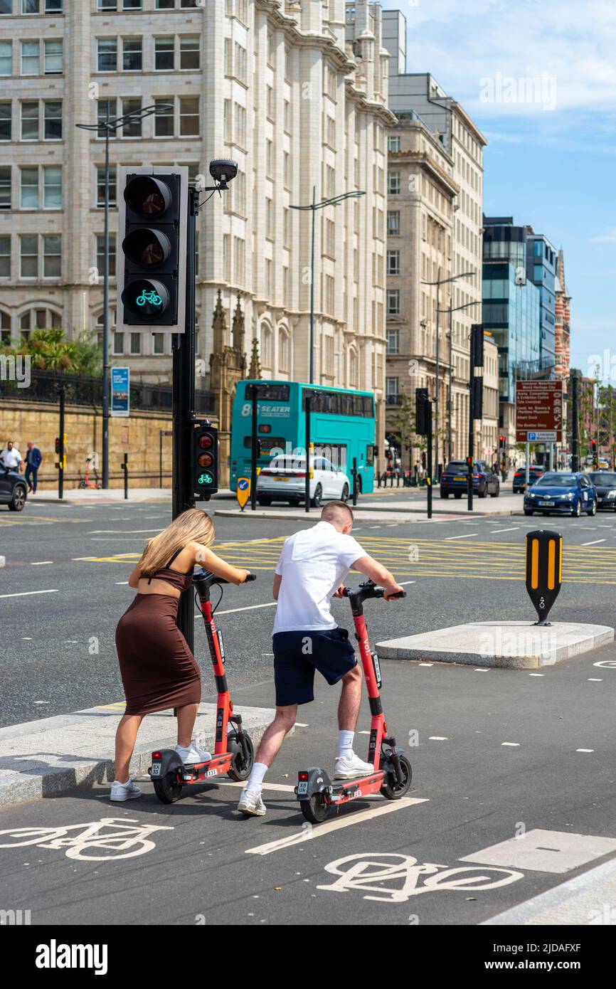 Giovane uomo e donna che cavalcano voi e-scooters che si spengono a un semaforo verde in una corsia per biciclette controllata al semaforo, Liverpool, Inghilterra, Regno Unito Foto Stock