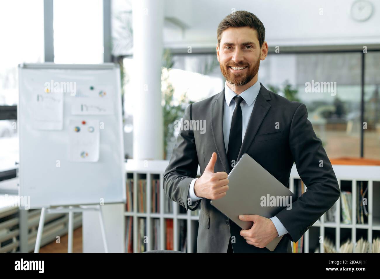 Elegante uomo d'affari caucasico positivo, ceo aziendale, auditor, si trova in un ufficio moderno, in una tuta aziendale formale, con un laptop nelle sue mani, guarda la fotocamera, sorride, mostra il gesto dei pollici Foto Stock