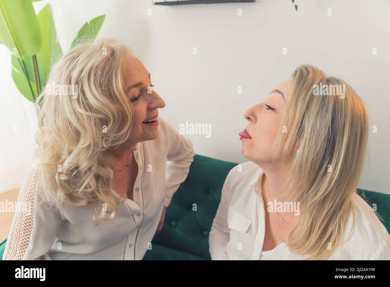 Due belle donne eleganti nel loro 40s seduta su un divano verde in un appartamento minimalista che si divertono e fare volti divertenti. Foto di alta qualità Foto Stock
