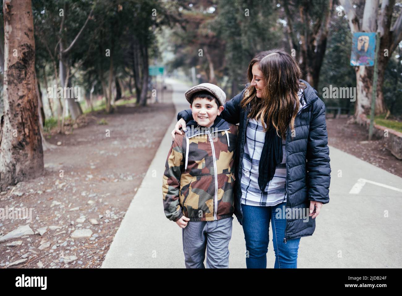 Giovane madre bionda che cammina e parla con suo figlio in una strada del parco. Famiglia con un solo genitore Foto Stock